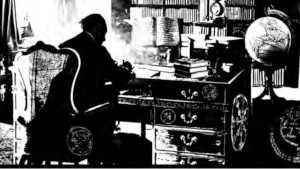 Un uomo, visto di schiena, è seduto alla scrivania di uno studio ricolmo di libri e di oggetti esoterici